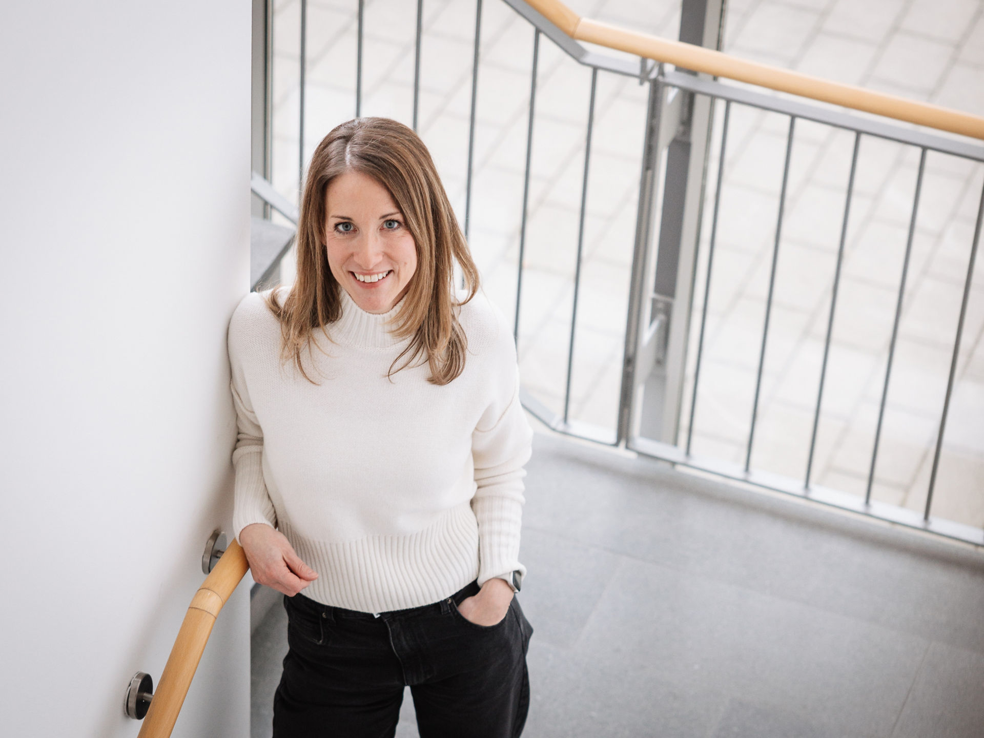 Frau mit hellbraunen Haaren trägt eine schwarze Hose und einen weißen Rollkragenpullover, lehnt an der Wand im Treppenhaus und lächelt hoch in die Kamera