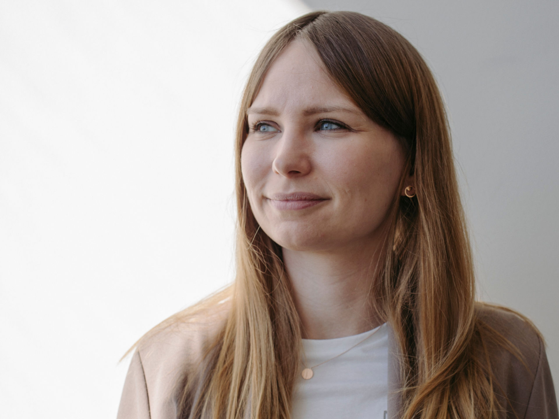 Frau mit hellbraunen Haaren steht vor einer weißen Wand und ihr Gesicht ist halb im Schatten. Sie trägt einen weißen T-shirt und beigen Blazer  und ihr Gesicht ist nach links gerichtet.
