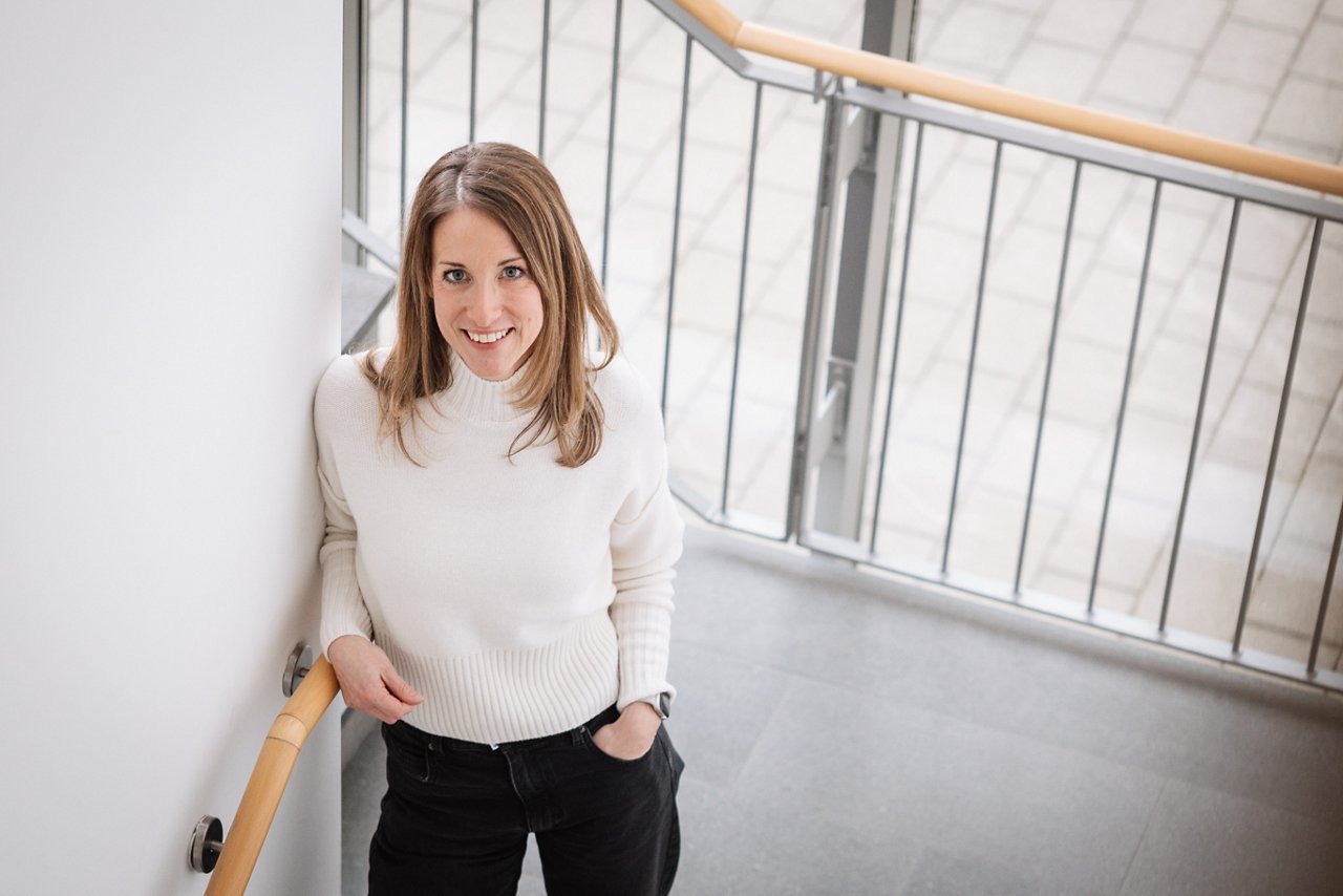 Woman with light brown hair wearing black pants and a white turtleneck, leaning against the wall in the stairwell and smiling high at the camera