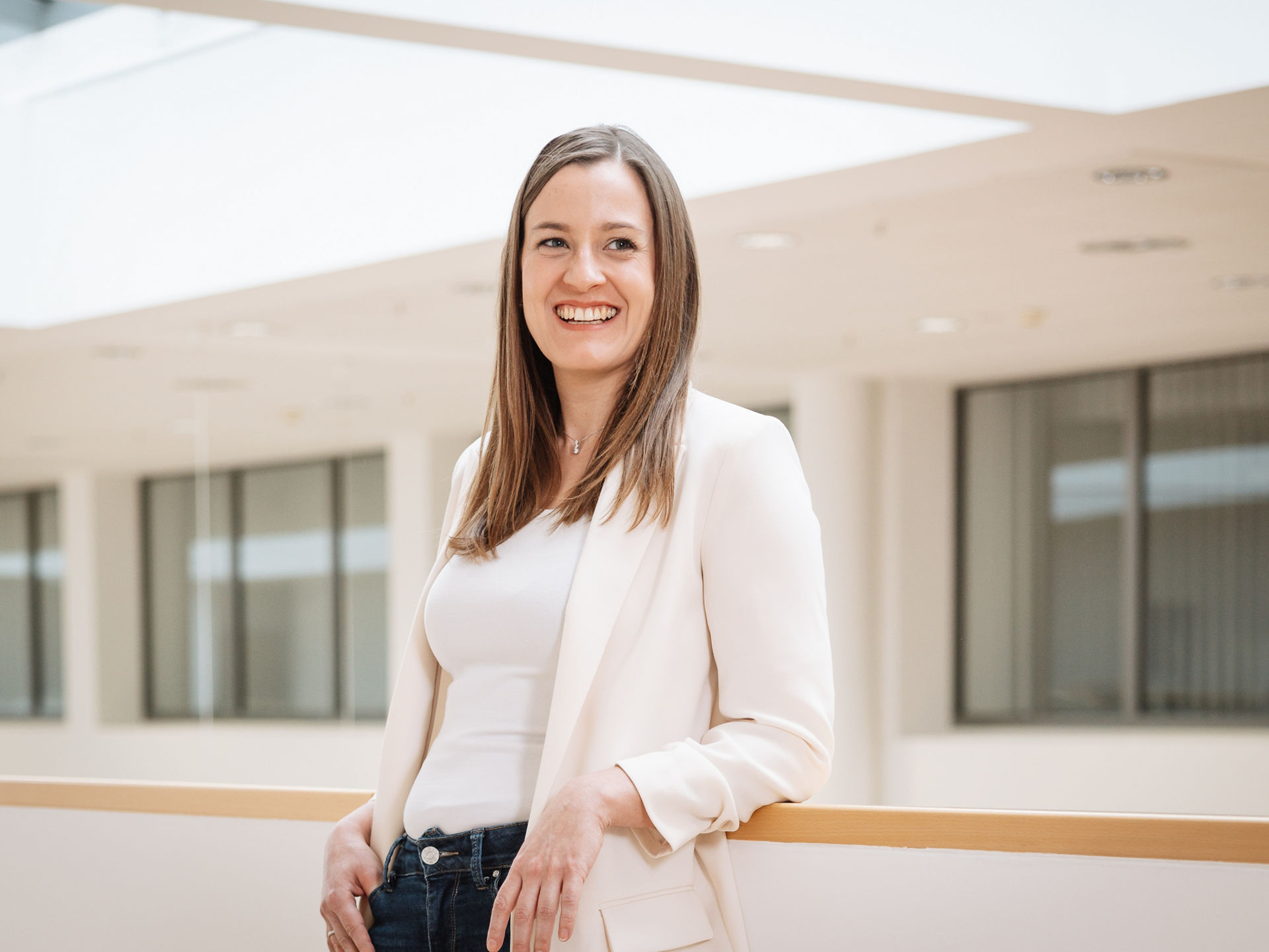 Frau mit hellbraunen, mittellangen Haaren trägt eine Jeans, ein weißes Tshirt und einen weißen Blazer. Sie stützt ihre Ellebogen auf ein halbhohes Geländer hinter sich und lächelt in die Kamera