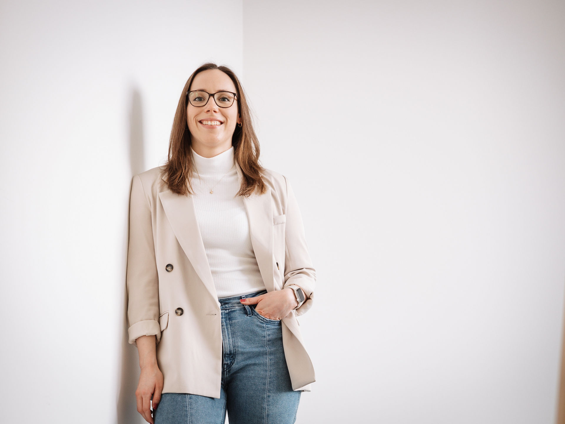 Braunhaarige Frau, Recruiterin bei Melitta, trägt eine Jeans, ein weißes Rollkragen-Shirt und beigen Blazer. Sie steht im Treppenhaus an die Wand gelehnt und lächelt in die Kamera
