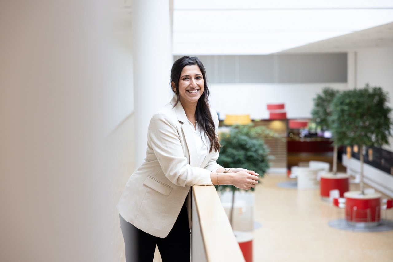 Dunkelhaarige Frau trägt einen weißen Blazer, lehnt über ein Geländer und lächelt