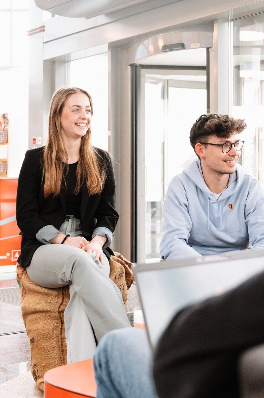 Two young colleagues sit on filled coffee bags in the entrance area and smile past the camera on the right. The young woman is wearing light-colored jeans and a black blazer. The young man to her right has brown hair and wears a light blue hooded sweater