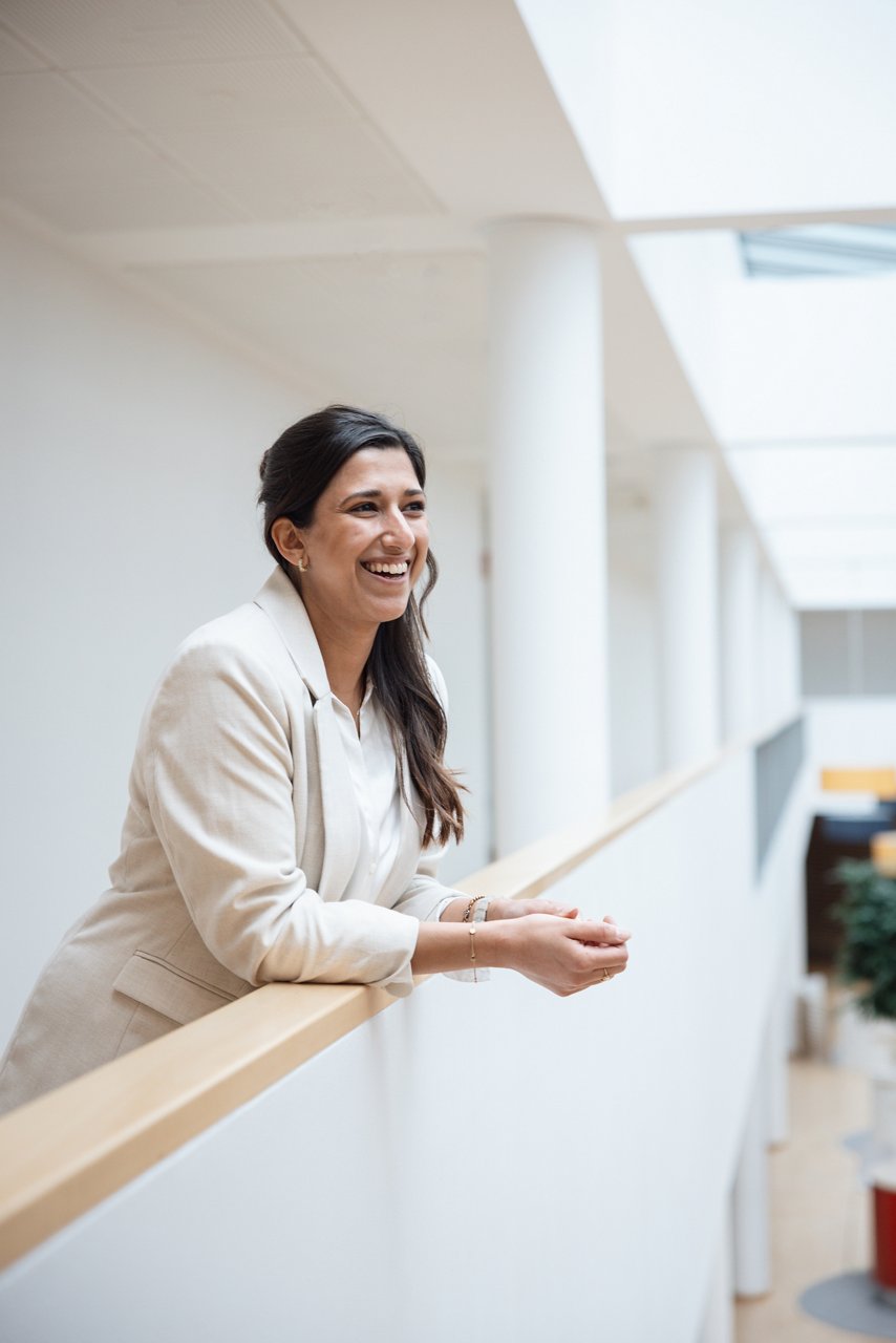 Brown-haired woman leaning with her arms on a half-height railing of a gallery. She wears a white blazer and smiles past the camera
