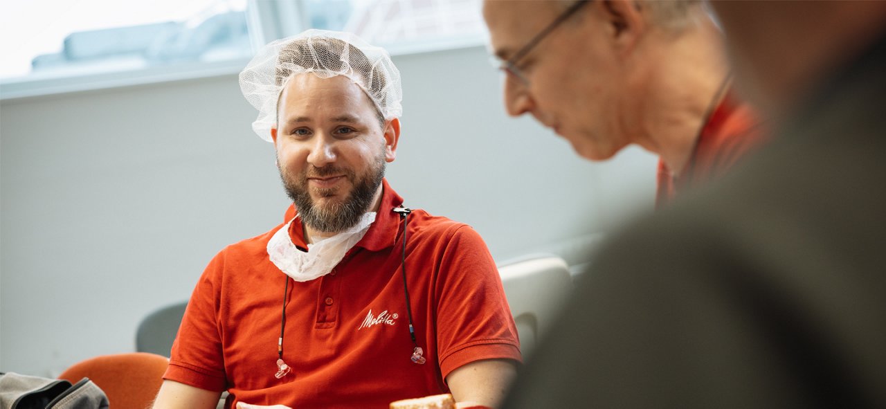 Melitta Mitarbeiter mit rotem Logotshirt und Produktionshaube auf dem Kopf spielen am Gemeinschaftstisch