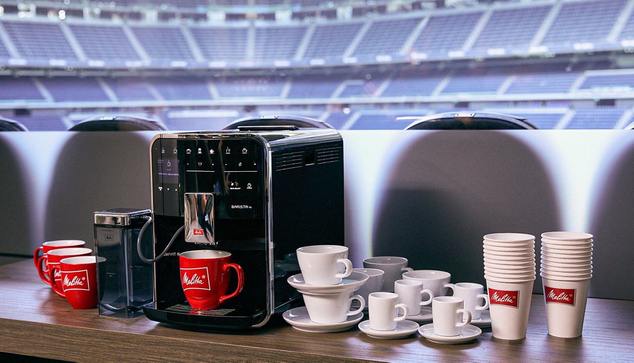 Eine schwarze Melitta Barista Kaffeemaschine steht auf einem Holztisch im Bernabéu-Stadion. Daneben sind Kaffeetassen, Untertassen und Melitta-Pappbecher aufgereiht. Im Hintergrund ist das Stadion zu sehen.
