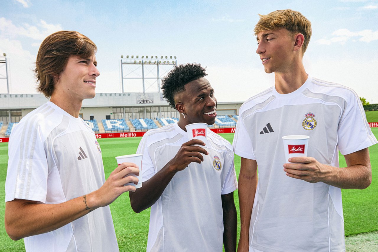 Drei Fußballspieler in weißen Real Madrid-Trikots stehen auf einem Trainingsplatz und halten Melitta-Kaffeebecher. Im Hintergrund sind Stadionsitze und Trainingsanlagen zu sehen.