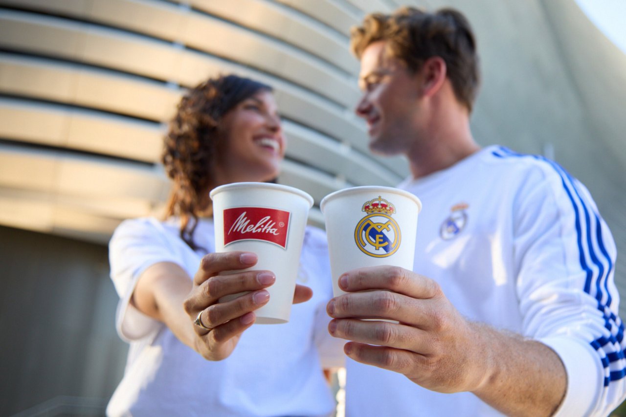 Ein junges Paar in weißen Trikots stößt mit Kaffeebecher an. Die Frau hält einen Becher mit Melitta-Logo, der Mann einen mit Real Madrid-Wappen. Beide lächeln sich an vor einer hellen Wand.