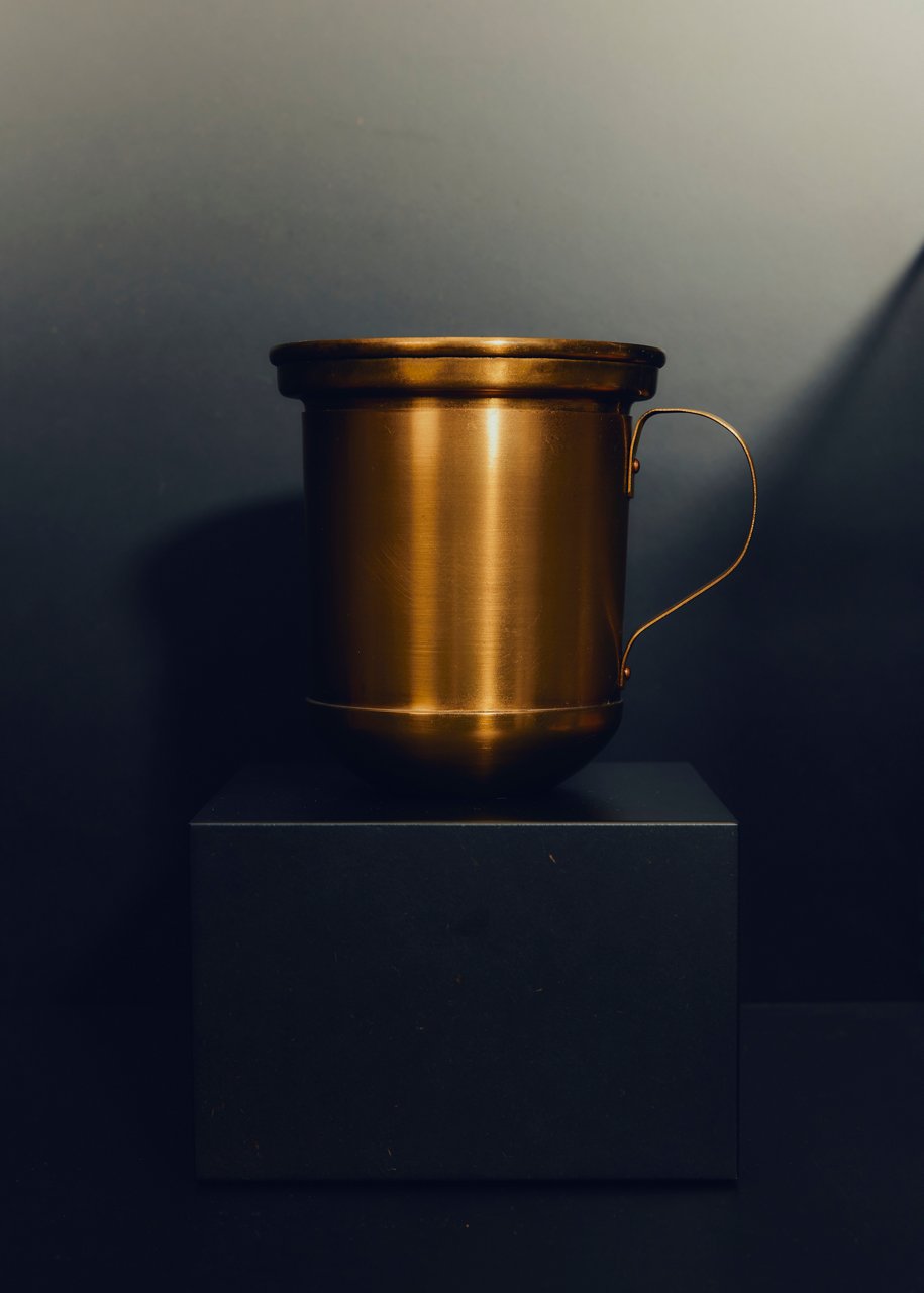 Goldfarbener Metallbecher mit Henkel, freigestellt auf einem schwarzen quadratischen Sockel vor dunklem Hintergrund, stimmungsvoll beleuchtet.