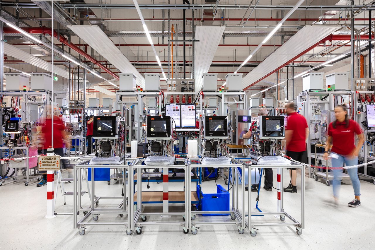 Moderne Produktionshalle mit einer Reihe von Fertigungsstationen für Kaffeevollautomaten; Mitarbeitende in roter Arbeitskleidung sind in Bewegung zwischen den Anlagen mit Bildschirmen und Rollwagen zu sehen.