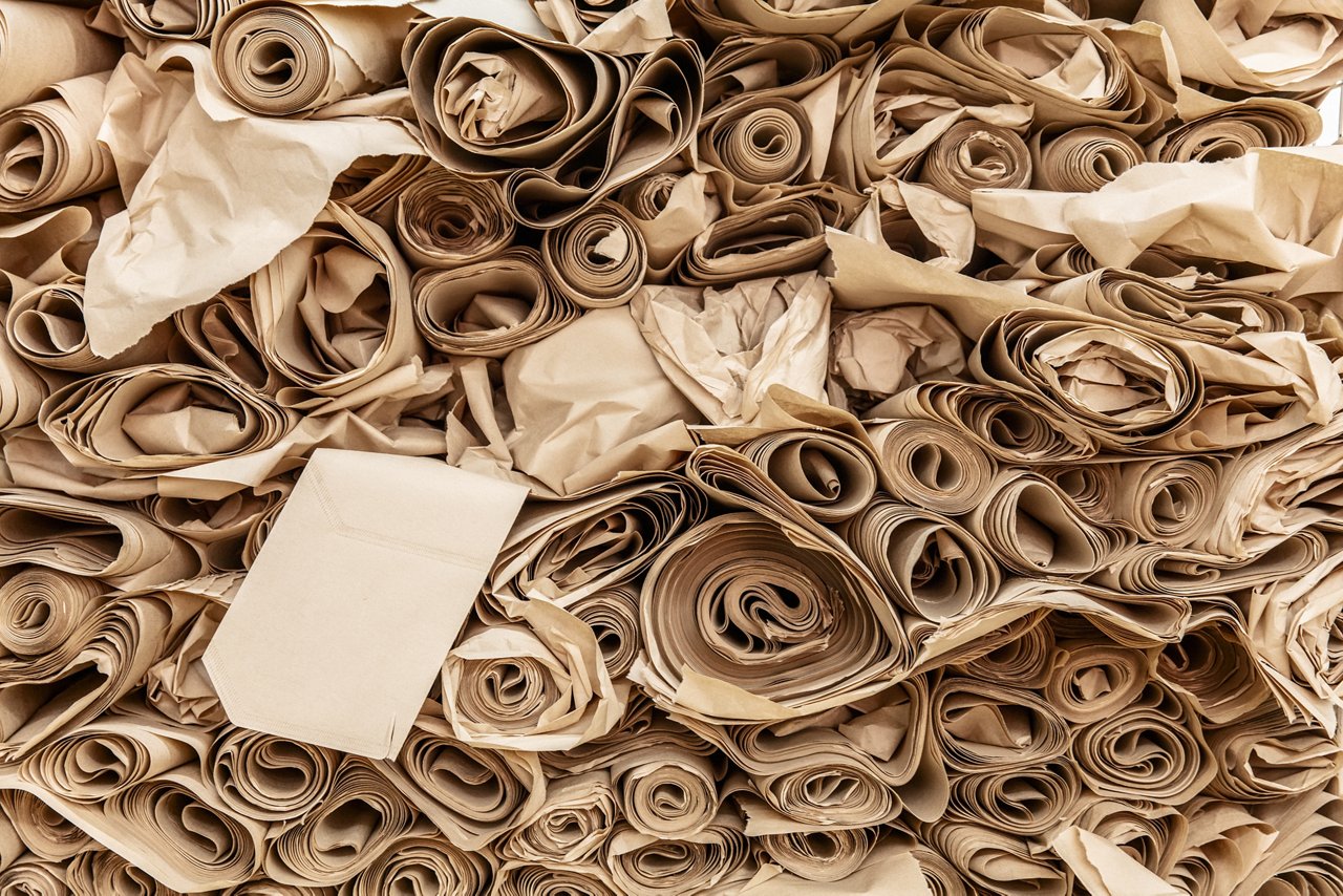 Draufsicht auf eine Vielzahl eingerollter und geknüllter brauner Papierstreifen und -bögen, die dicht aneinanderliegen; mittig ein flach liegender heller Papierumschlag.