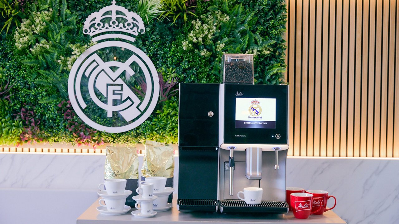 Eine Melitta XT6 Kaffeemaschine steht auf einer Marmorplatte vor einer grünen Pflanzenwand mit dem silbernen Real Madrid-Logo. Daneben stehen Kaffeetassen und Kaffeepakete mit der Aufschrift "Bacio d'Oro".