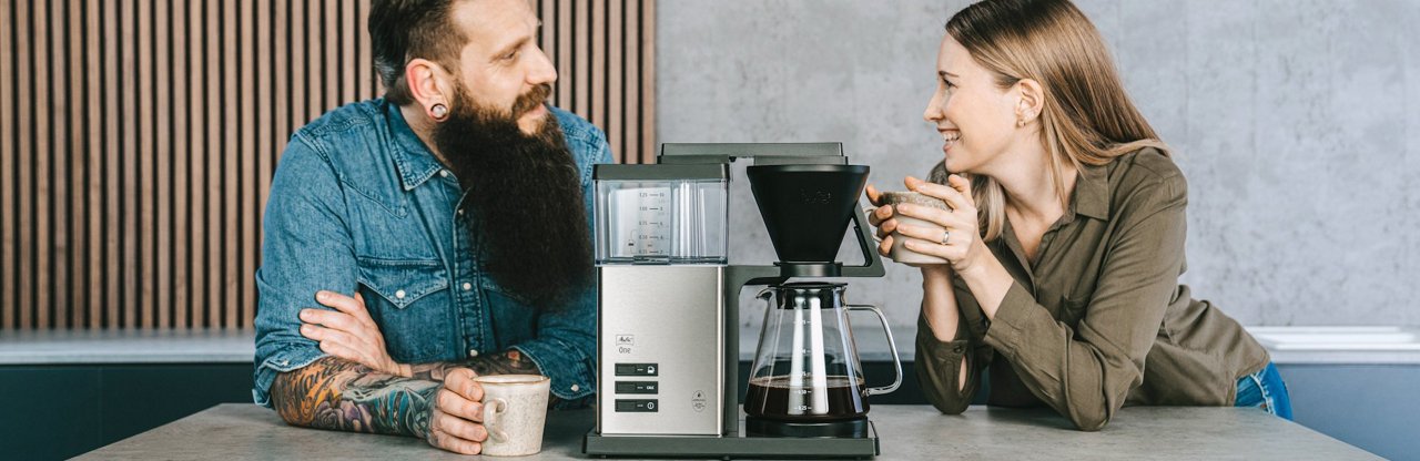 Melitta® One Filter Coffee Maker placed in the center of a kitchen board with two people leaning around and chatting both holding coffee mugs in their hands