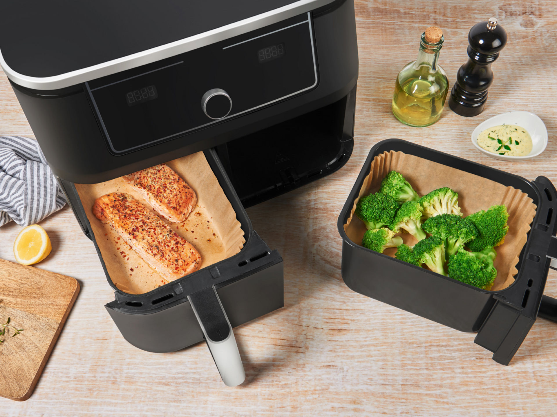 Fächer einer Heißluftfritteuse mit Backpapier ausgelegt: Im linken Fach liegen zwei gewürzte Lachsfilets, im rechten Fach frische Brokkoliröschen. Auf dem Tisch liegen Zitronen, Kräuter, Öl, Pfeffermühle und eine Schale mit Dip.
