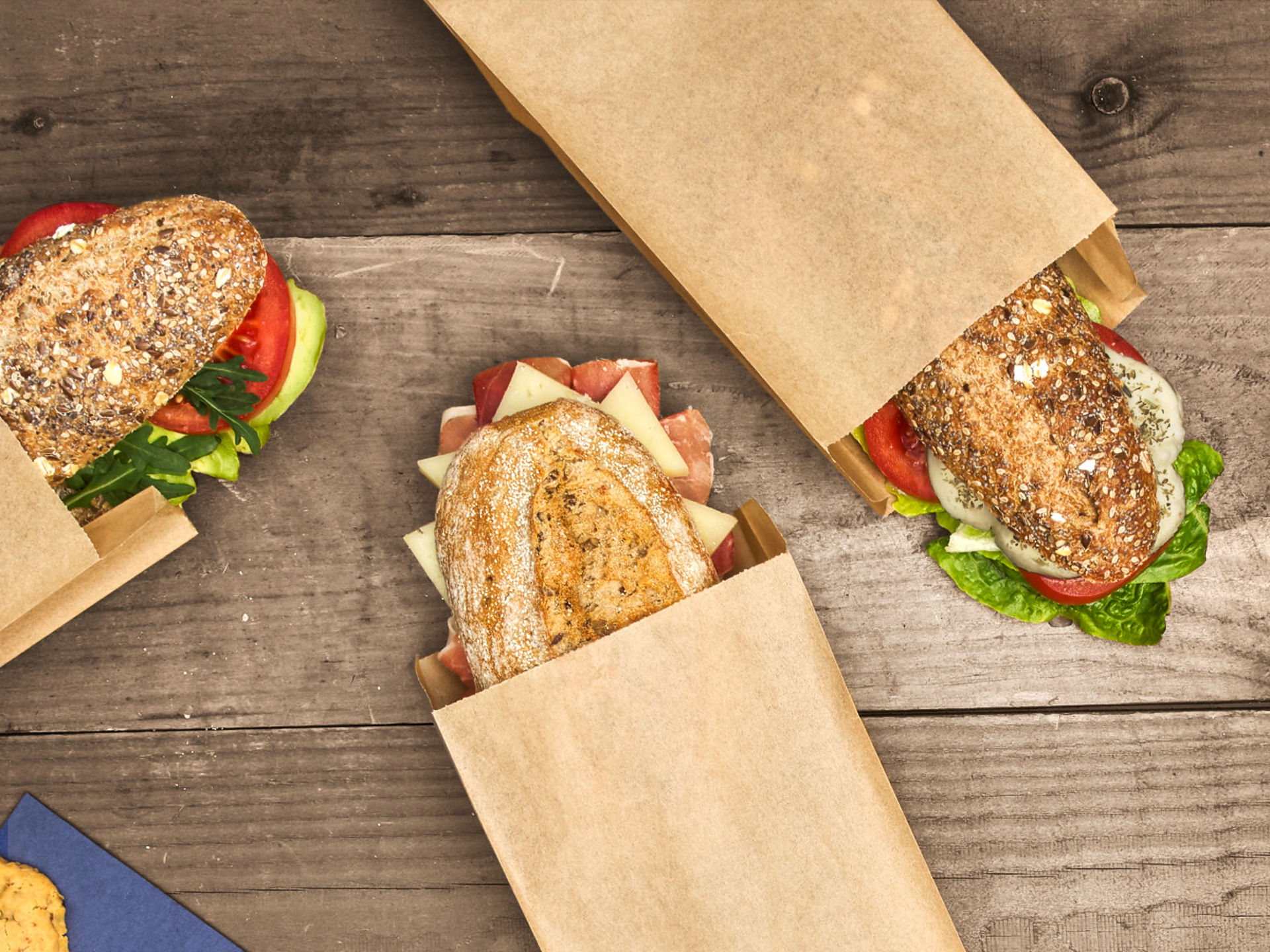 Drei belegte Brötchen in Sandwich-Papiertüten liegen auf einem Holztisch. 