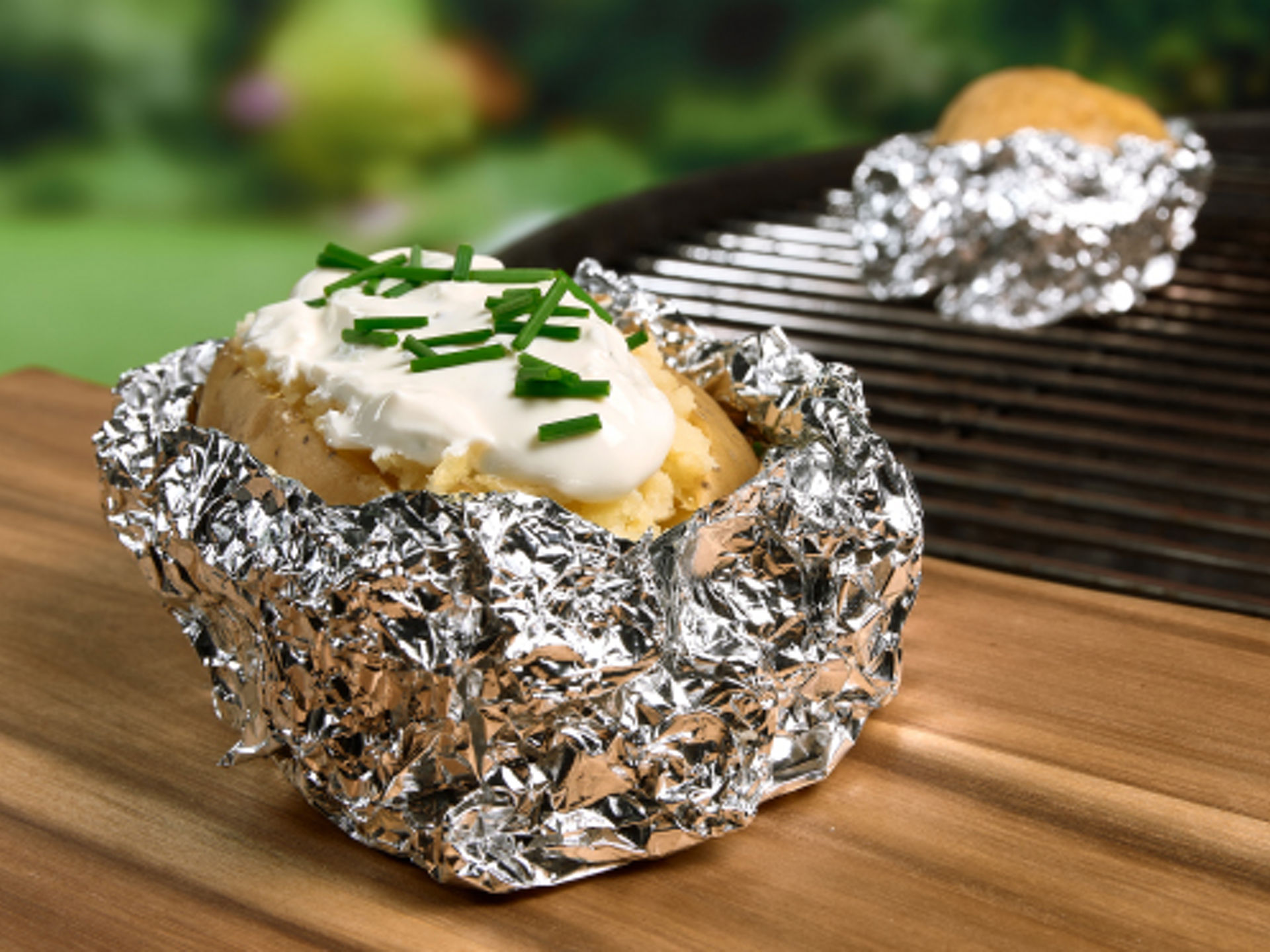 En bagekartoffel indpakket i Toppits® aluminiumsfolie med cremefraiche og purløg ligger på et træbord, og en grill ses i baggrunden