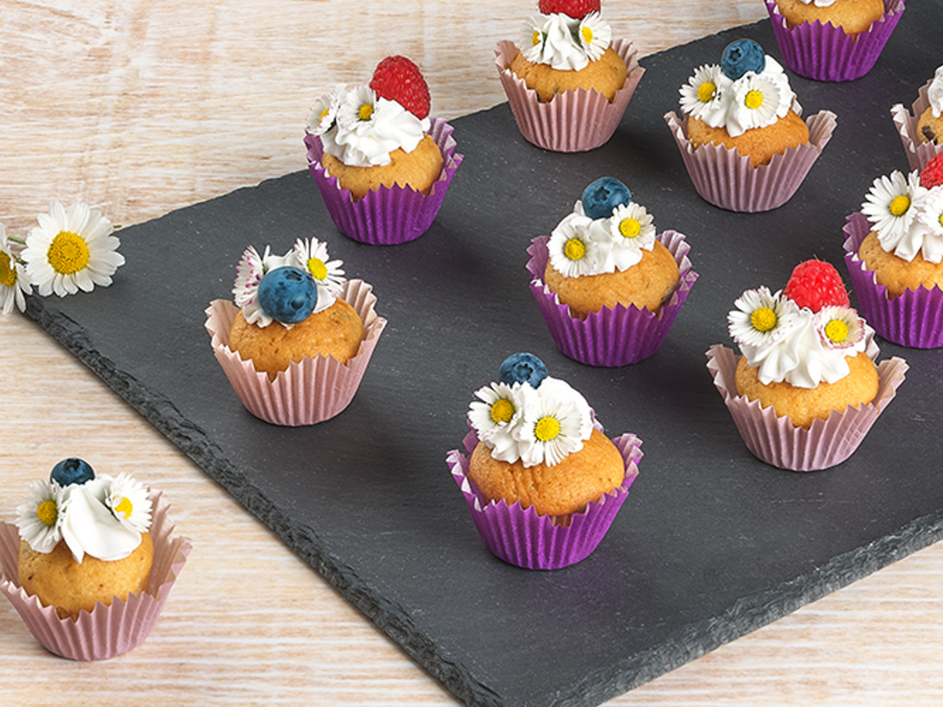 Kleine Muffins in bunten Toppits  Papierförmchen, verziert mit Sahne, essbaren Gänseblümchen, Blaubeeren und Himbeeren, auf einer schwarzen Schieferplatte angerichtet 