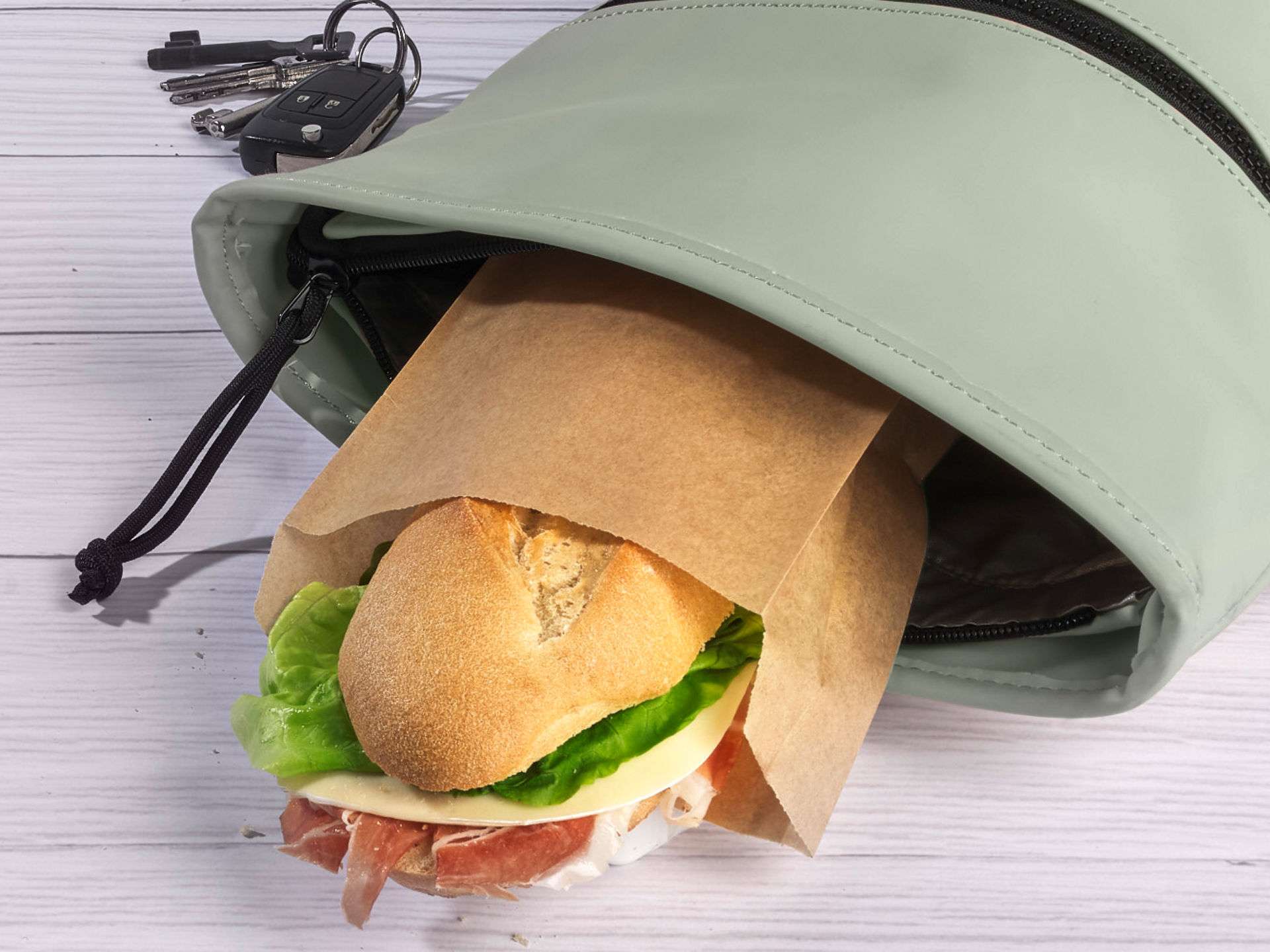 Ein belegtes Brötchen in einer Sandwich-Papiertüte steckt in einer geöffneten Handtasche, daneben liegen Schlüssel. 