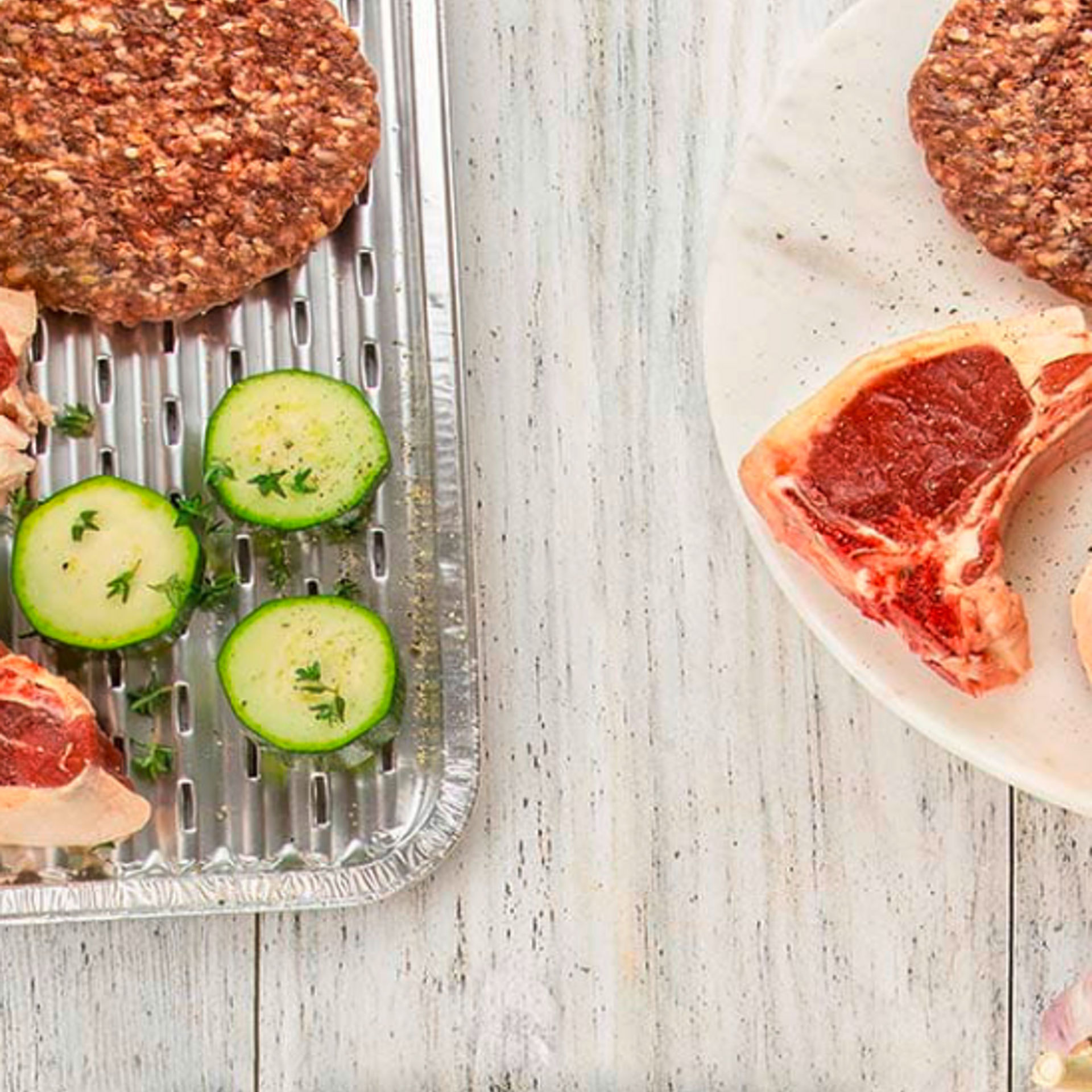 Eine Toppits Express Grillpfanne mit rohem Fleisch, Burger-Patties, Mais und Zucchinischeiben, daneben ein Teller mit weiteren Fleischstücken und Burger-Patties auf einem hellen Holztisch