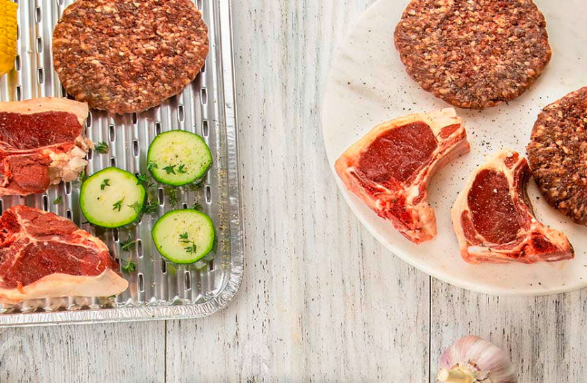 Eine Toppits Express Grillpfanne mit rohem Fleisch, Burger-Patties, Mais und Zucchinischeiben, daneben ein Teller mit weiteren Fleischstücken und Burger-Patties auf einem hellen Holztisch