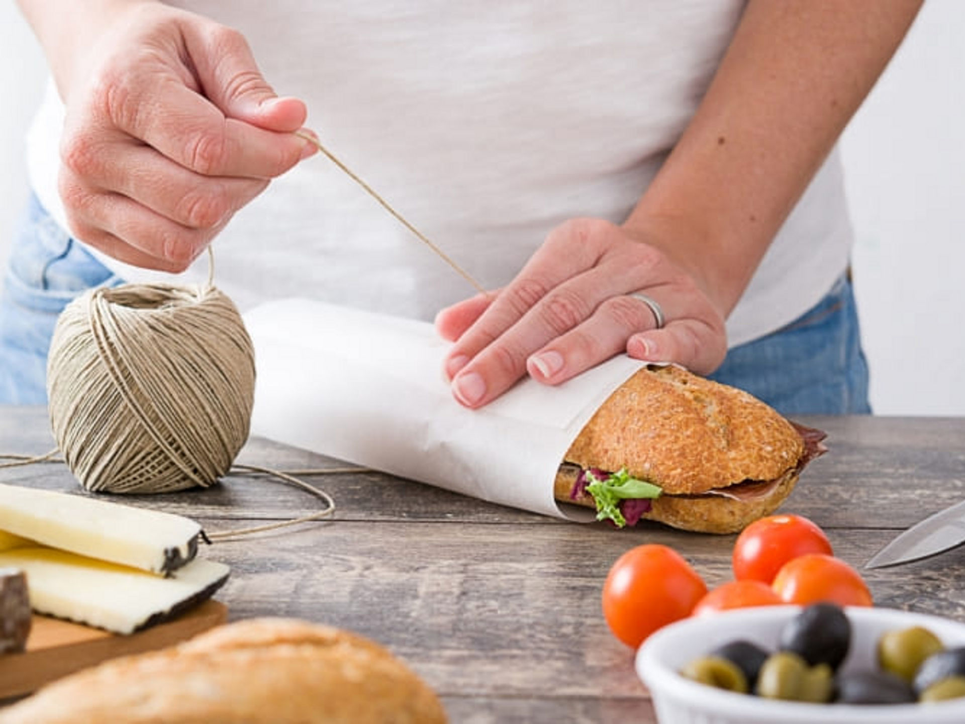 Eine Person wickelt ein belegtes Baguette in Toppits Butterbrotpapier und bindet es mit einer Schnur, daneben liegen Käse, Tomaten und Oliven auf dem Tisch