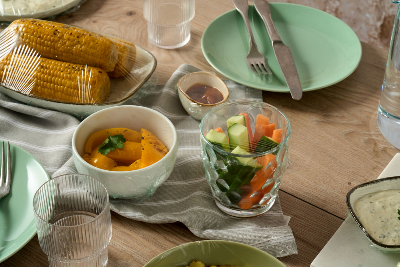 Bord med forskjellige retter og en glassbeholder dekket med Toppits plastfolie.