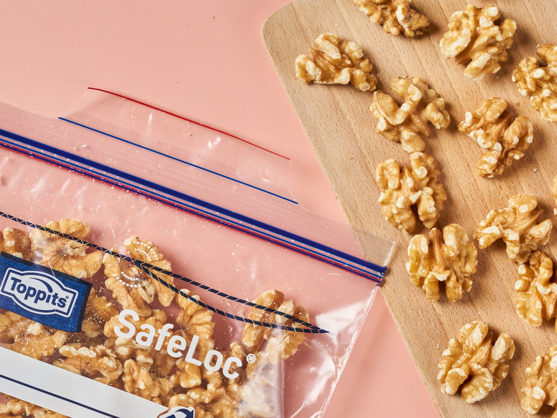 Toppits SafeLoc Beutel gefüllt mit Walnusskernen liegt neben Holzbrett auf rosa Fläche.