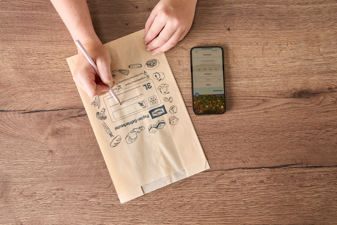 Eine Person beschriftet einen Toppits Papier-Gefrierbeutel auf einem Holztisch neben einem Smartphone.