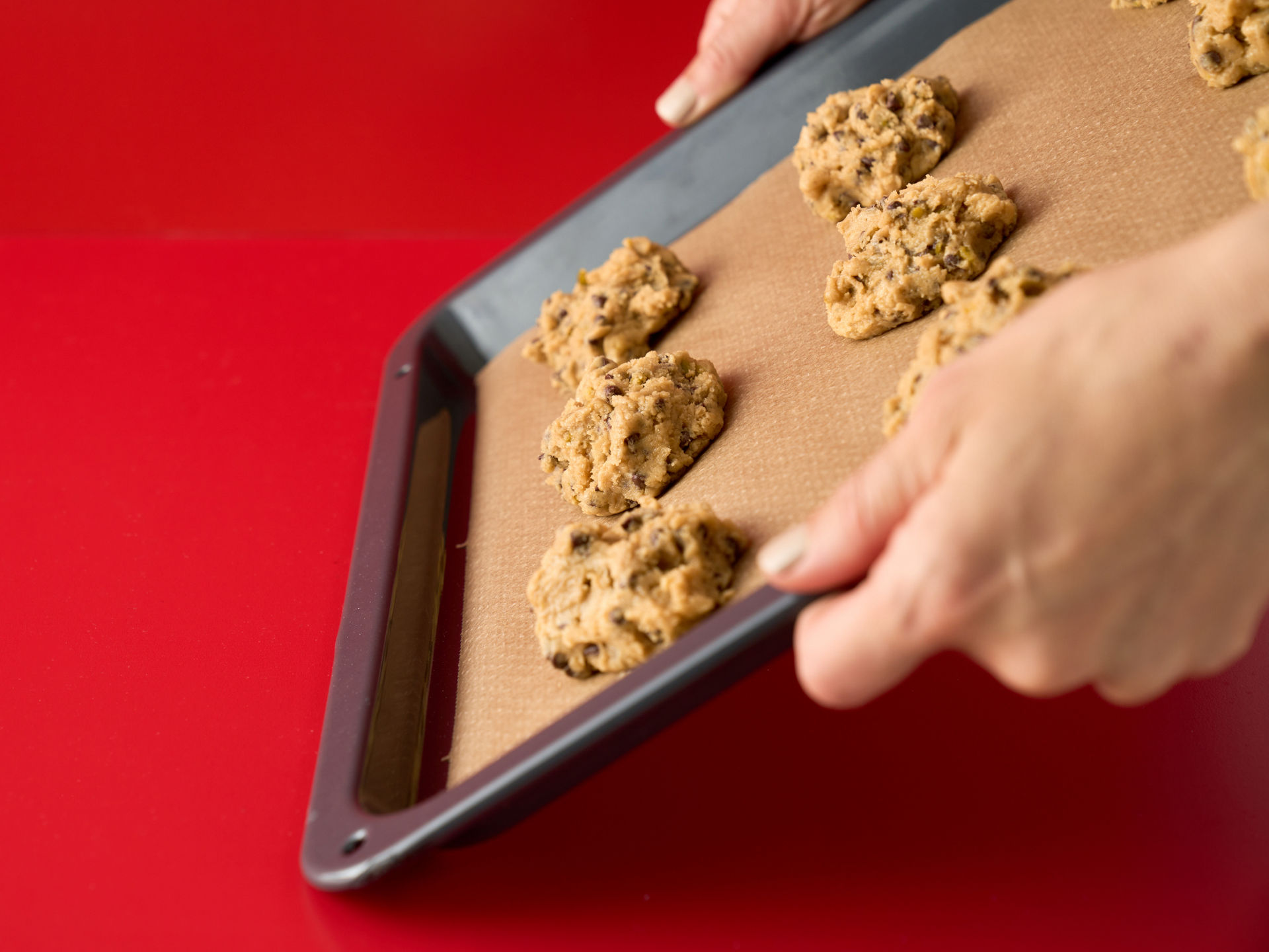 Hände halten ein Backblech mit Keksteig auf Toppits Backpapier über rotem Hintergrund.