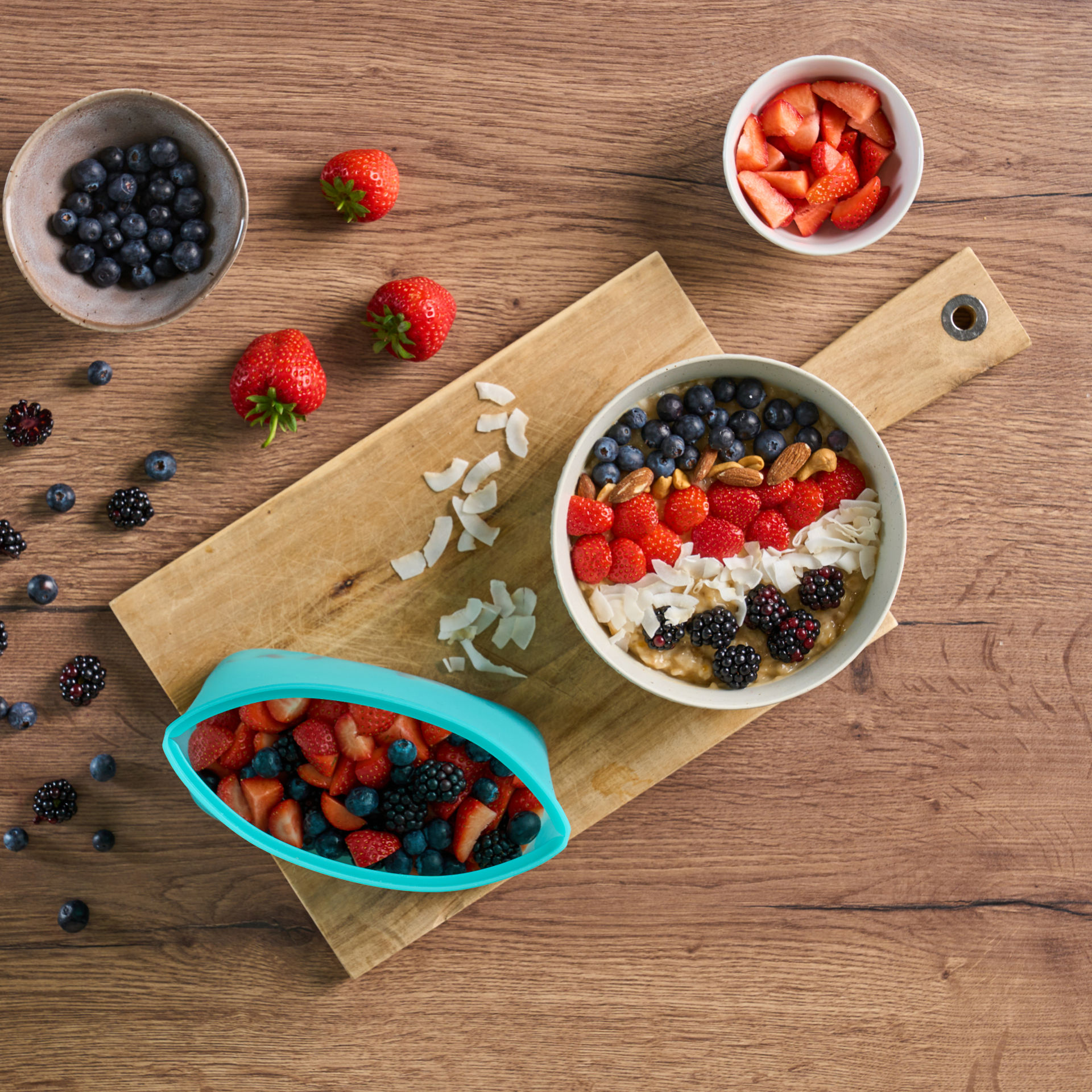 Frühstückszubereitung mit frischen Beeren, Nüssen und Haferbrei auf einem Holzbrett mit Silikonbeutel.