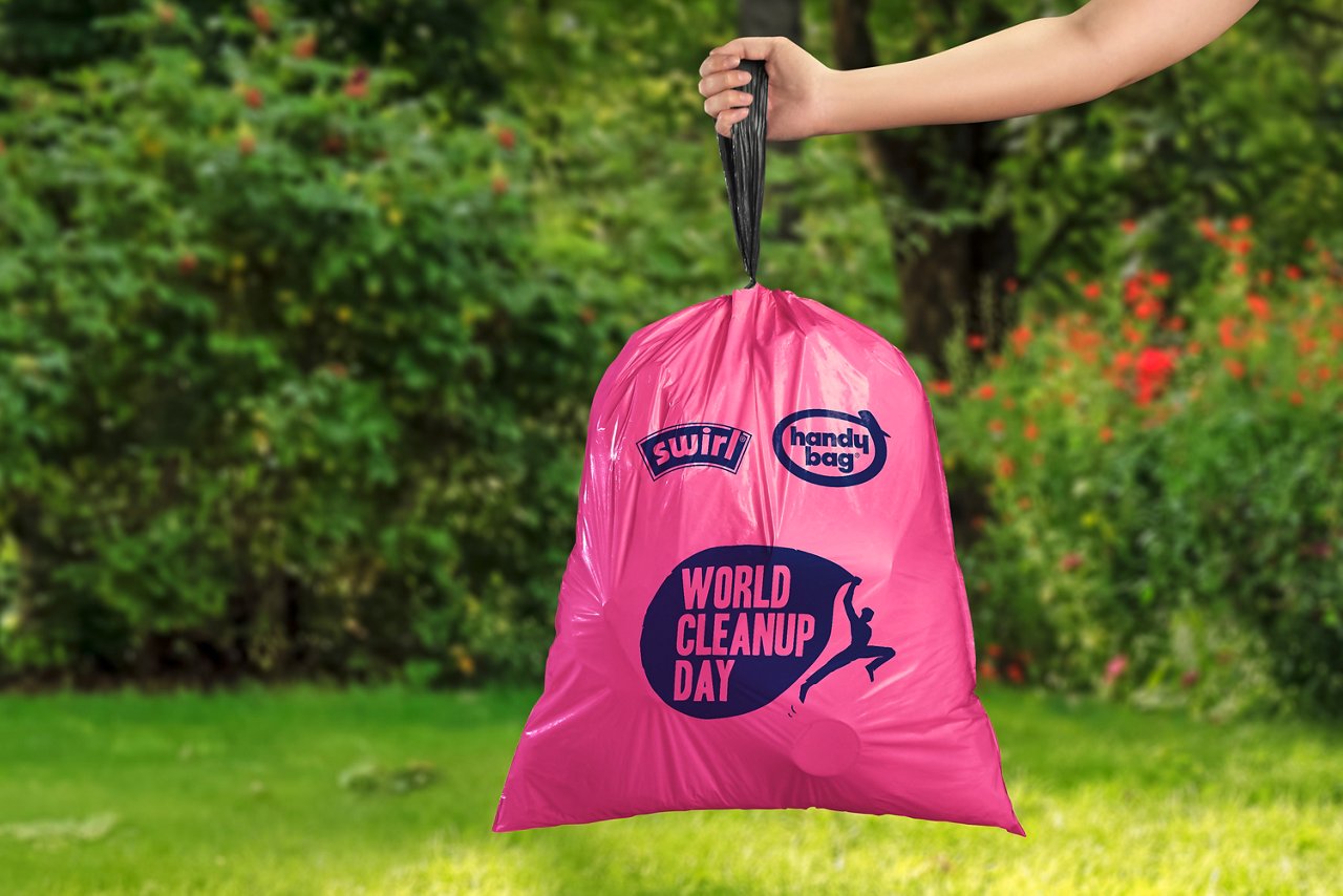 Ein pinker Swirl Handy Bag vollen Müllbeutel mit World Cleanup Day Aufdruck wird im Garten an einer Hand hochgehalten