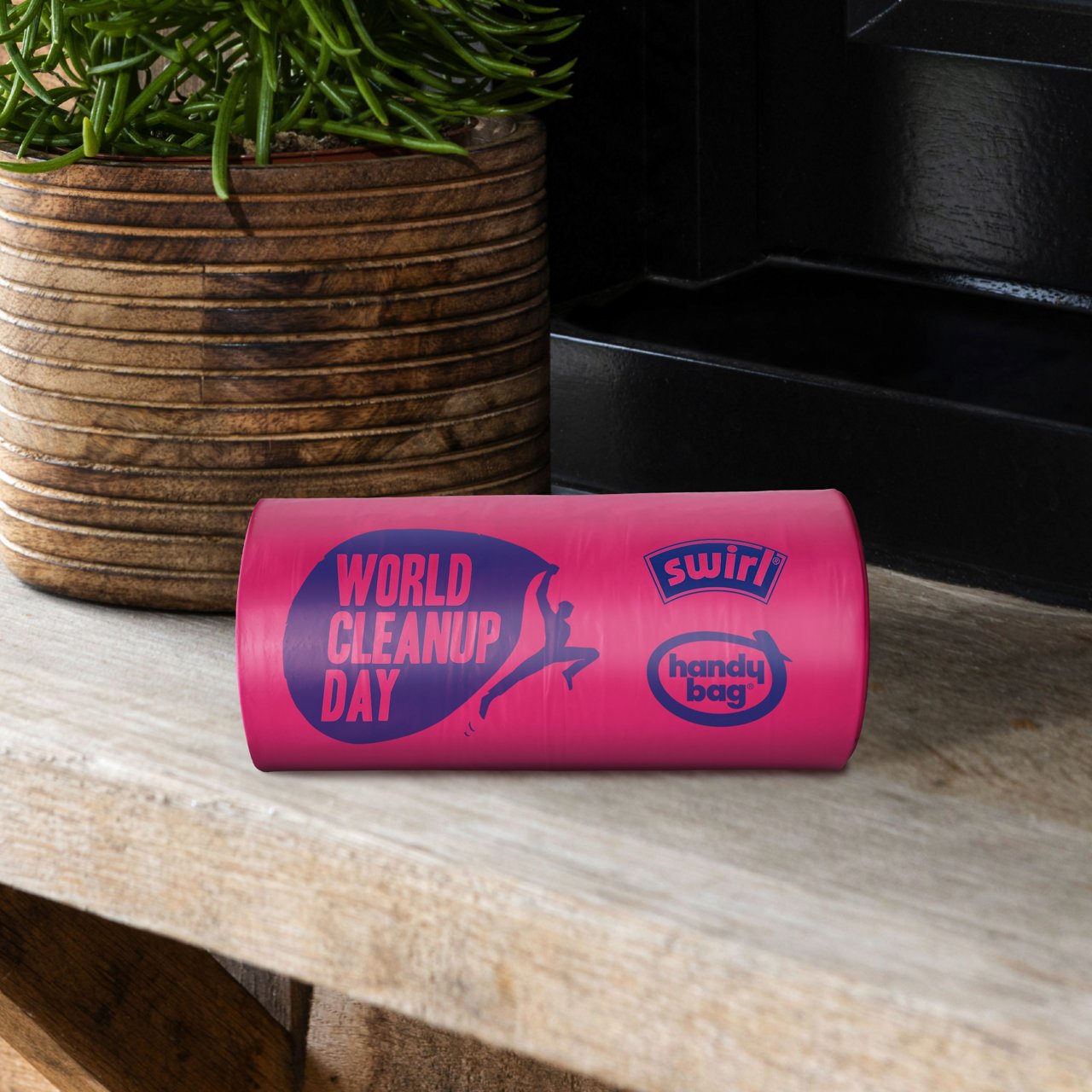 Eine pinke Swirl Handy Bag Müllbeutel Rolle mit World Cleanup Day Branding liegt auf einer Holzbank neben einer Pflanze