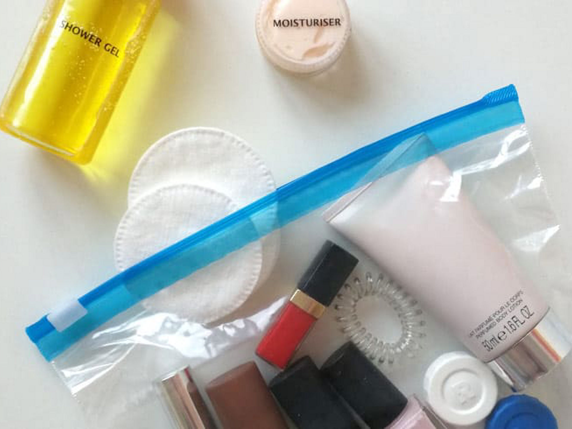 Transparenter Toppits Zipper-Beutel mit verschiedenen Kosmetikartikeln wie Creme, Lippenstift, Nagellack und Haargummi, daneben liegen Wattepads, ein gelbes Duschgel und ein kleiner Tiegel mit Feuchtigkeitscreme auf weißem Untergrund  