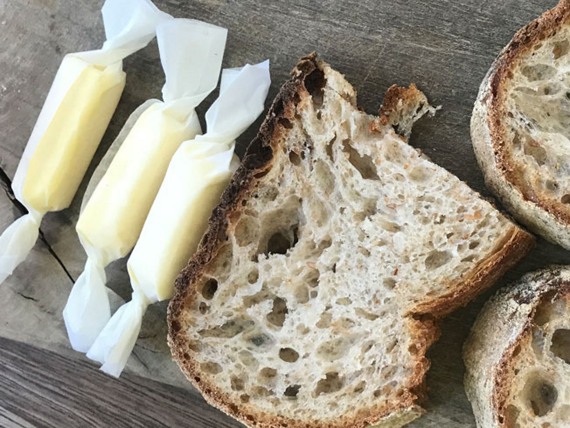 Drei Scheiben Bauernbrot und drei in Toppits Butterbrotpapier gewickelte Butterstücke liegen auf einem Holztisch