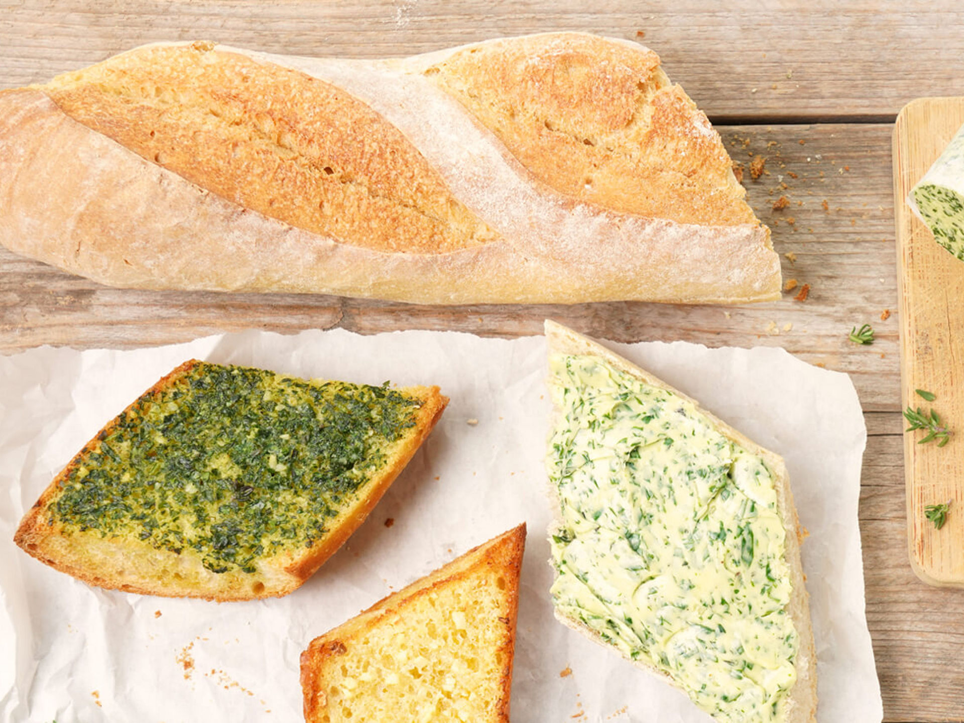 Ein Baguette, mehrere Scheiben Knoblauchbrot und Kräuterbutter liegen auf einem Holzbrett. Auf einem weiteren Brett sind zwei Rollen Kräuterbutter, ein Messer und eine Knoblauchzehe zu sehen