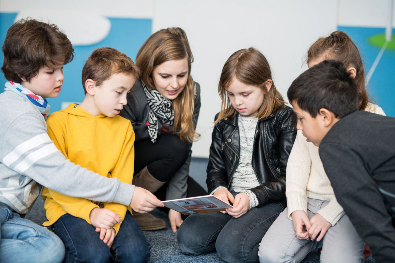 Mehrere Kinder sitzen in einer Gruppe auf dem Boden und schauen gemeinsam auf ein Blatt Papier, das sie in den Händen halten. Im Hintergrund ist eine Wand mit bunten Elementen zu sehen