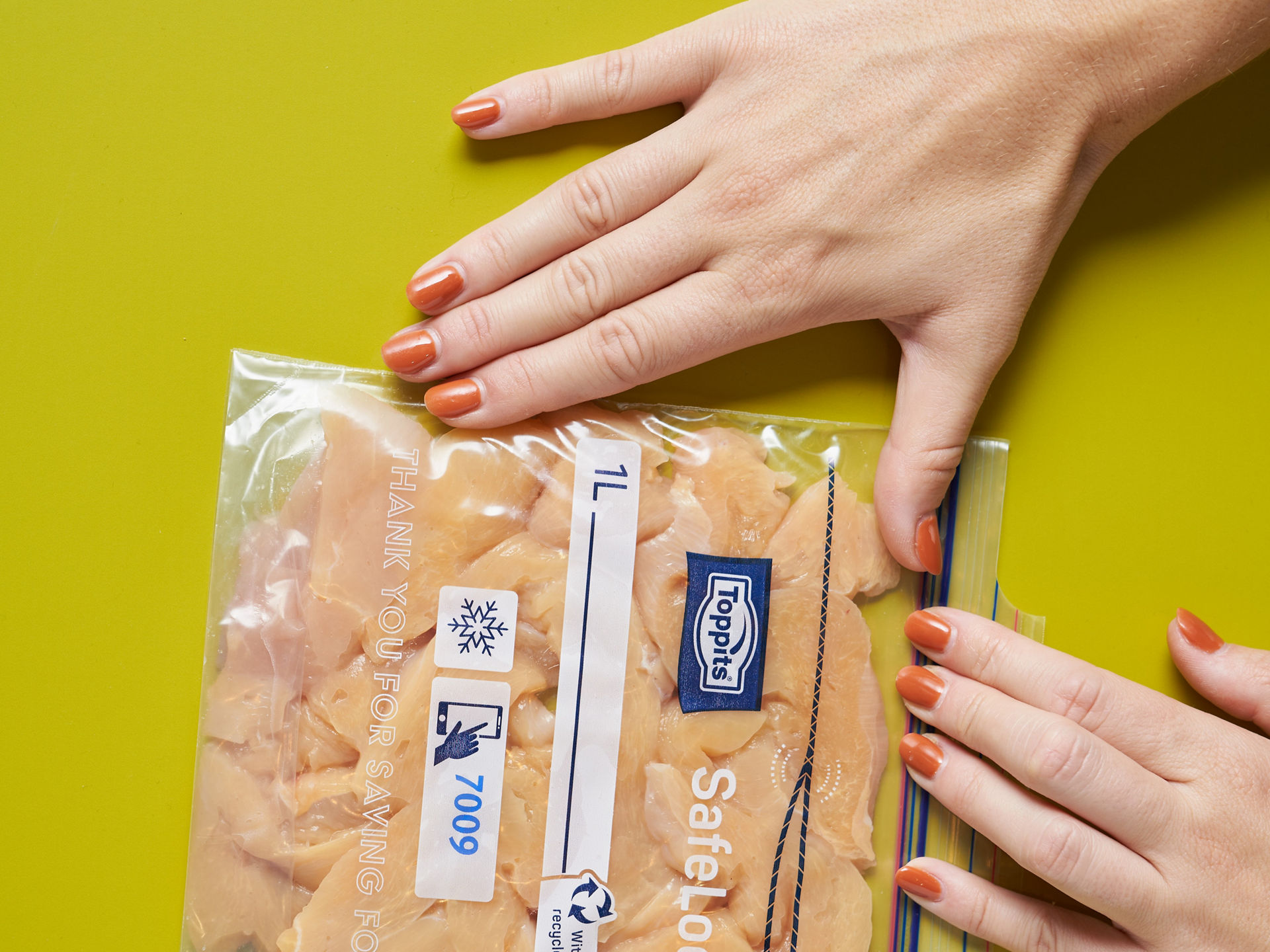 Zwei Hände drücken einen Toppits SafeLoc Gefrierbeutel mit rohem Hähnchenfleisch auf einer gelben Fläche flach