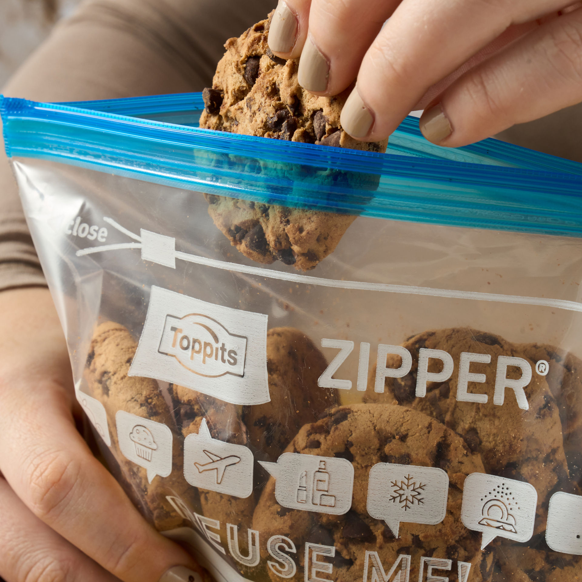 Eine Hand nimmt einen Schoko-Cookie aus einem Toppits Zipper Beutel auf einem Holztisch mit gelbem Tuch.