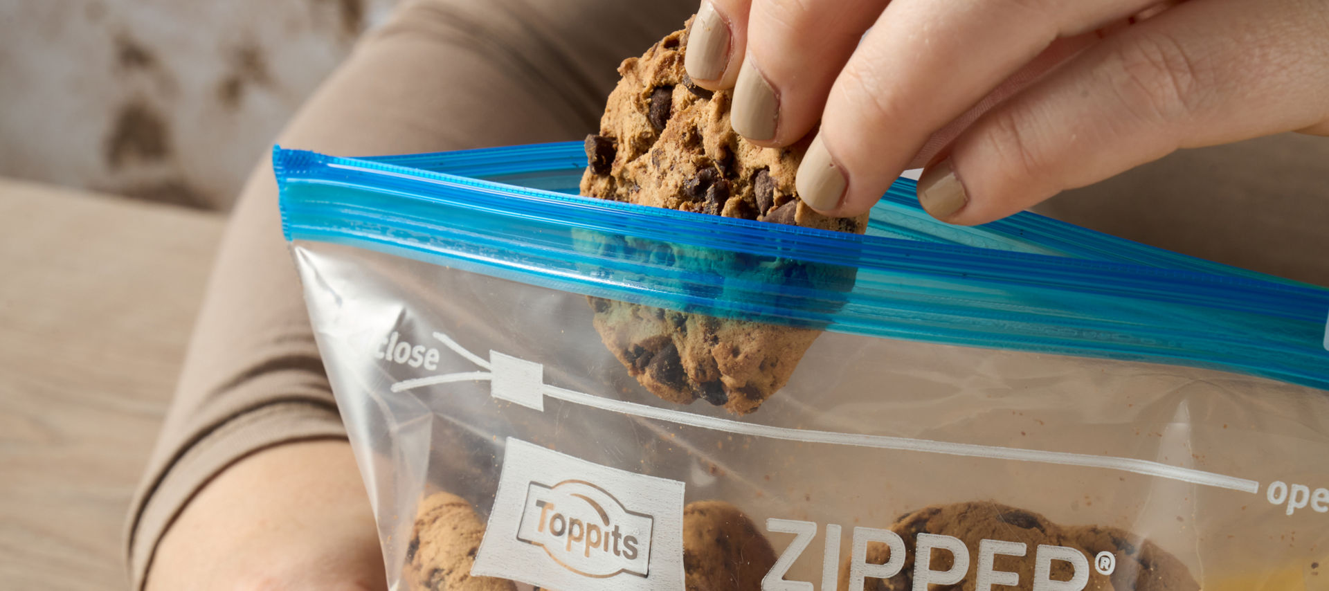 Eine Hand nimmt einen Schoko-Cookie aus einem Toppits Zipper Beutel auf einem Holztisch mit gelbem Tuch.
