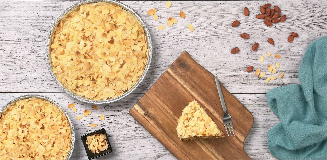 Bakad mandeltårta på träbräda, mandlar och mandelspån på ljust träbord.