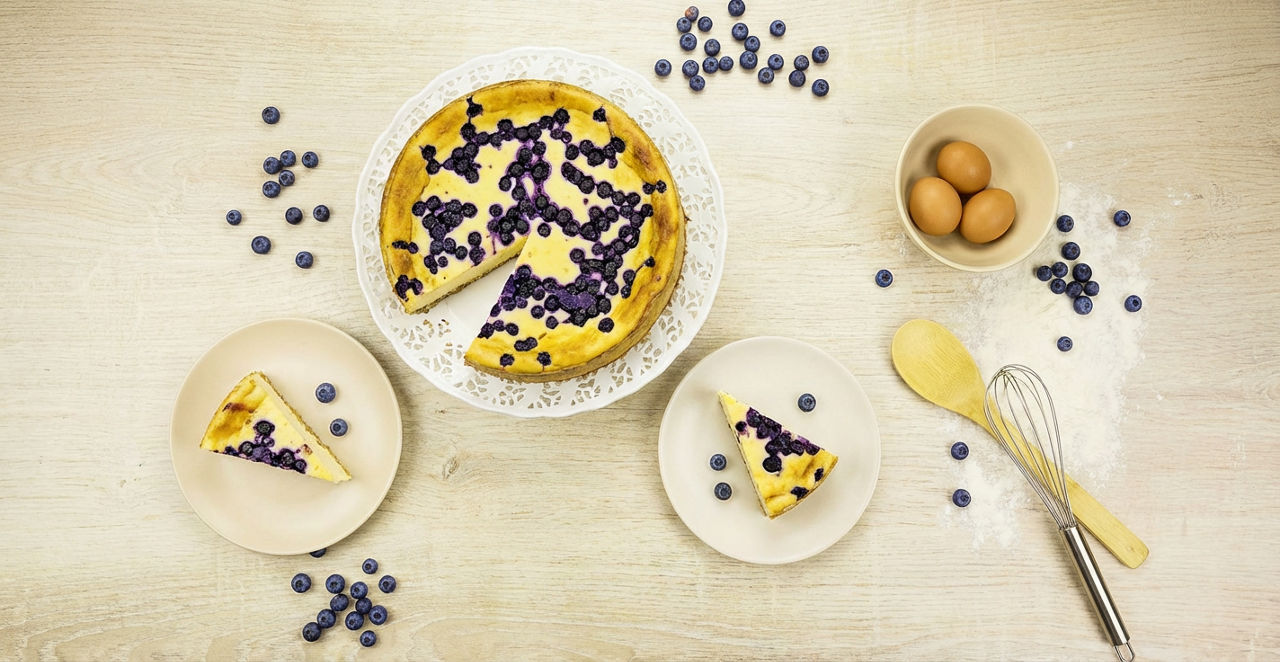 Bakad blåbärskaka, en tallrik med tårtbit, ägg, blåbär och bakredskap på ett ljust bord.