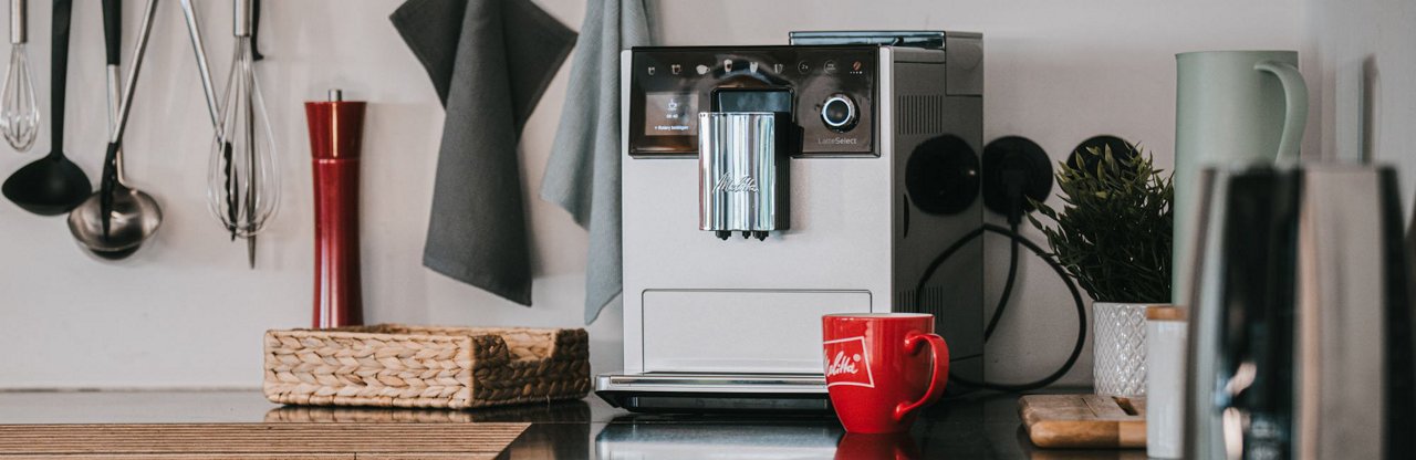 fam_latteselect_2000x650px.jpg – Melitta® LatteSelect coffee machine in studio setting with modern design and red cup placed beneath the dual spout.