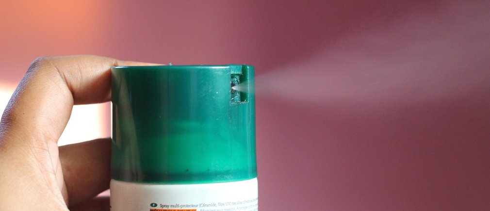 Mano sosteniendo un aerosol verde que libera una fina bruma sobre un fondo rosa desenfocado