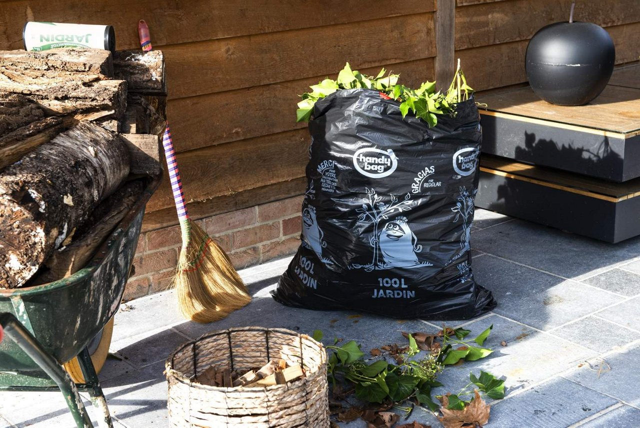 Sac poubelle Handybag jardin 100 litres rempli de feuilles vertes à côté d’une brouette