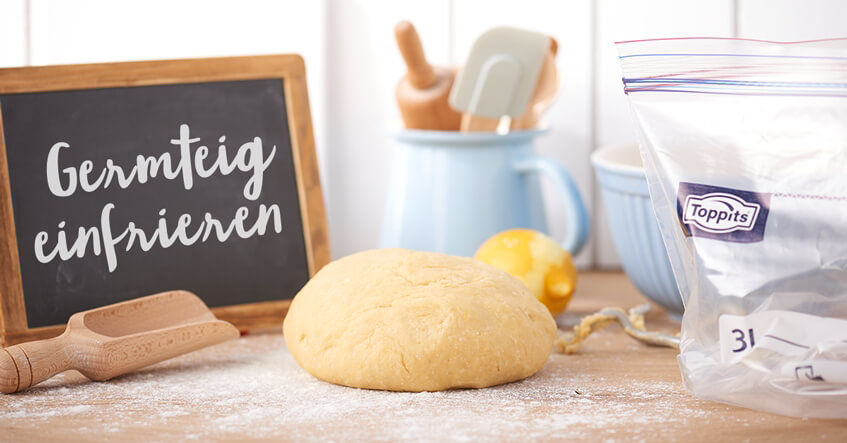 Germteig liegt auf einer bemehlten Arbeitsfläche, daneben ein Toppits® Gefrierbeutel und eine kleine Tafel mit der Aufschrift „Germteig einfrieren“. Im Hintergrund stehen Backutensilien und eine Zitrone.