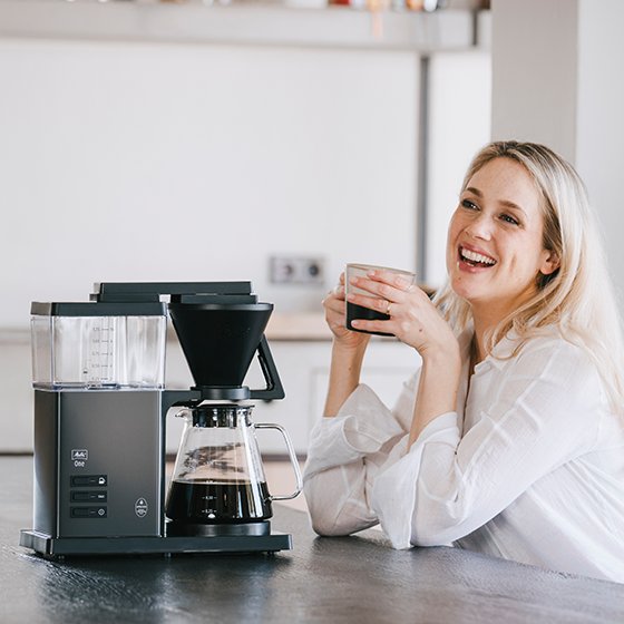 Melitta® One Filter Coffee Maker 6780186 placed on kitchen table with woman beside smiling and holding a coffee mug
