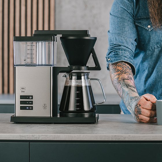 Melitta® One Filter Coffee Maker 6780188 front view coffee machine standing on kitchen board coffee jug filled with coffee man standing beside the coffee machine holding a coffee mug