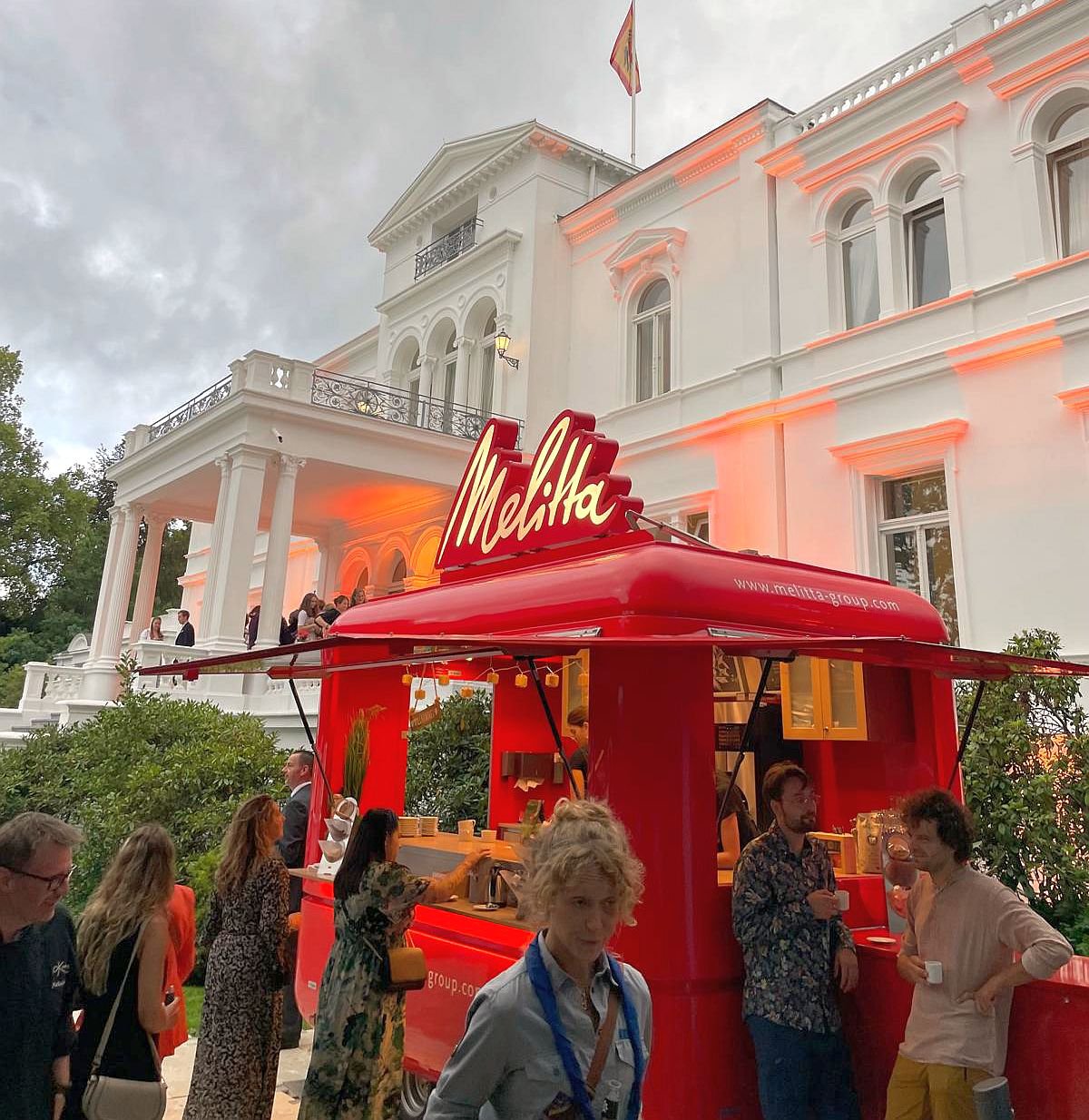 Der rote Melitta-Kaffeewagen mit goldenem Schriftzug steht abends vor der orange beleuchteten Villa Hammerschmidt, umgeben von Gästen.