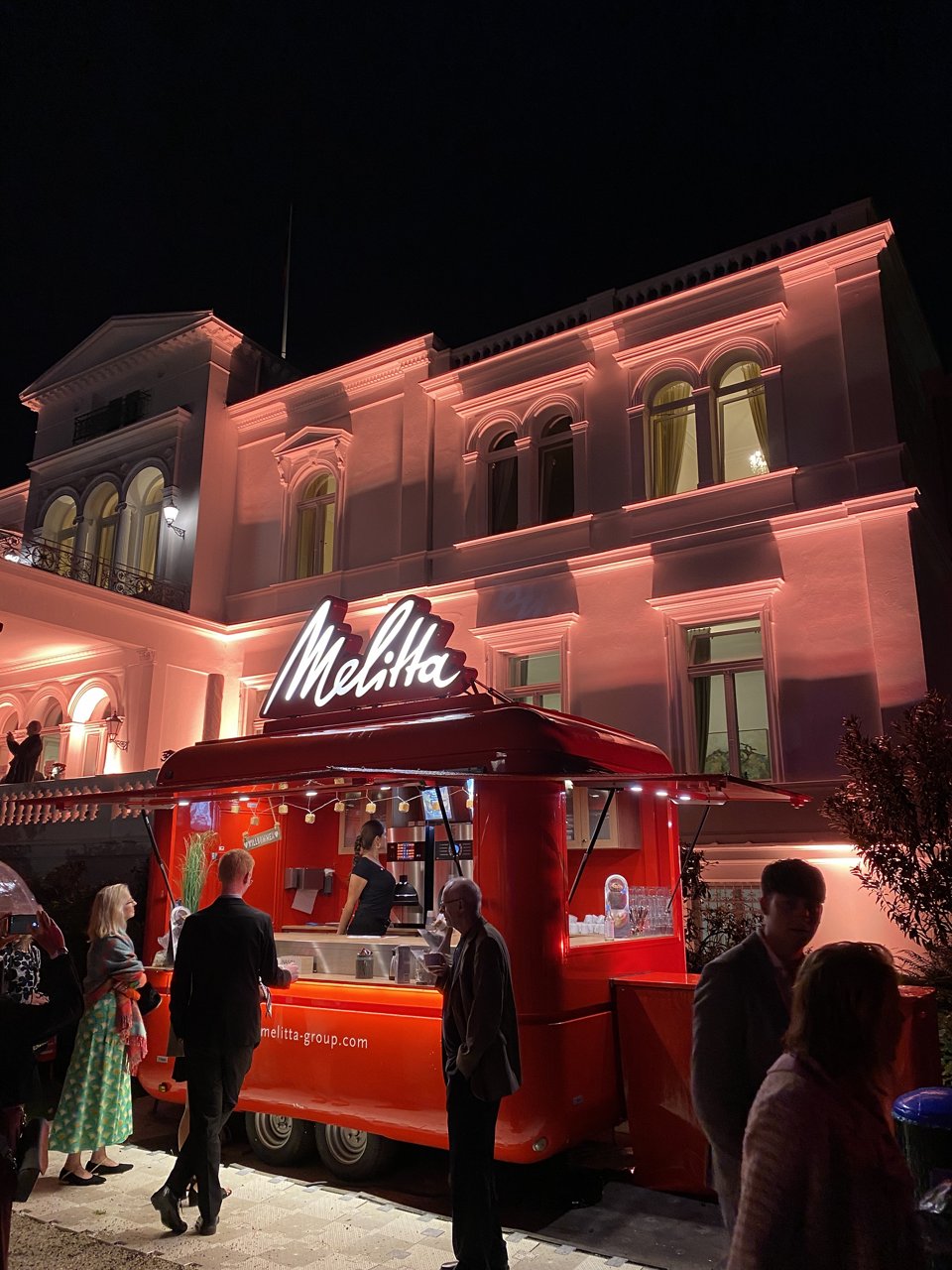 Nachts beleuchteter roter Melitta-Kaffeewagen mit weiß leuchtendem Melitta-Schriftzug vor einem orange angestrahlten historischen Gebäude, davor mehrere Personen.