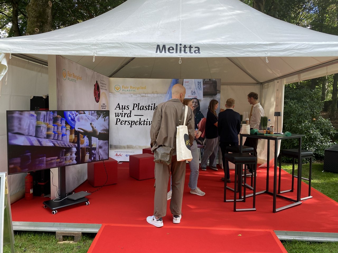Im Melitta-Pavillon steht ein Besucher vor einem Banner mit der Aufschrift „Aus Plastik wird – Perspektive" und einem Bildschirm, der den Recyclingprozess von Plastik zeigt.